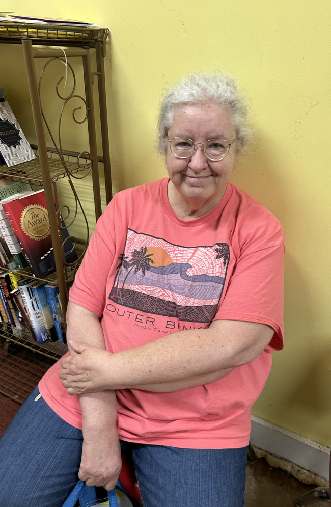 An older woman with curly white hair and glasses smiles while seated near a bookshelf.