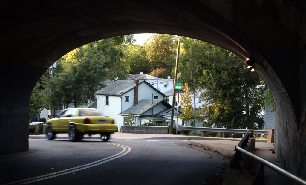 A road runs underneath the darkened arch of a bridge and curves just after the bridge ends. A lifted car with large tires has just driven under the bridge and zips around the curve. In the background is a dense collection of homes.