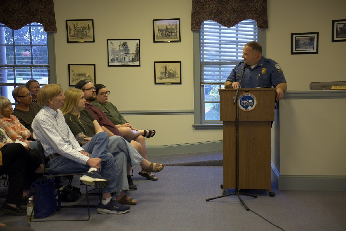 A man in a blue police uniform stands at a wooden podium and speaks to a small crowd of seated people to his side. There are windows and black and framed black and white drawings of buildings on the wall behind them.