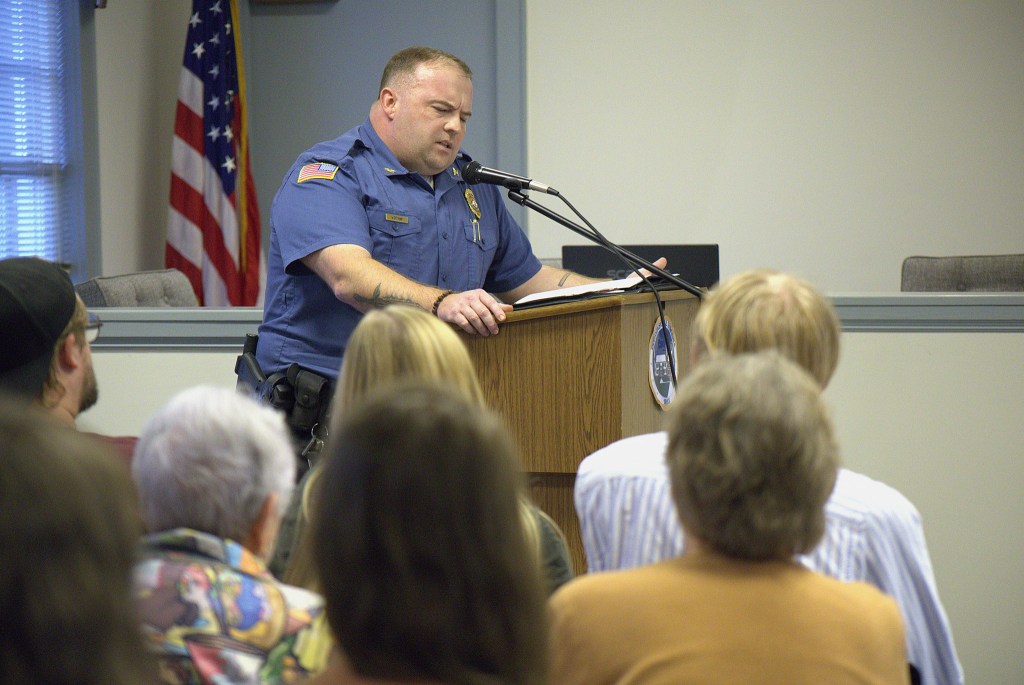 A man in a blue police uniform stands at a wooden podium and speaks into a microphone. He is looking down and reading from a folder with papers resting on the podium. A small crowd is seated in the foreground, watching the police officer and facing away from the camera.