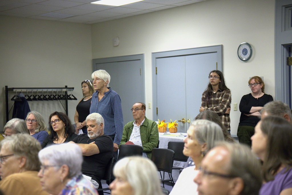 A small crowd of people sit in a meeting room with white walls and gray doors. Several people stand in the back of the room. They are looking forward and listening intently to someone who is off camera.