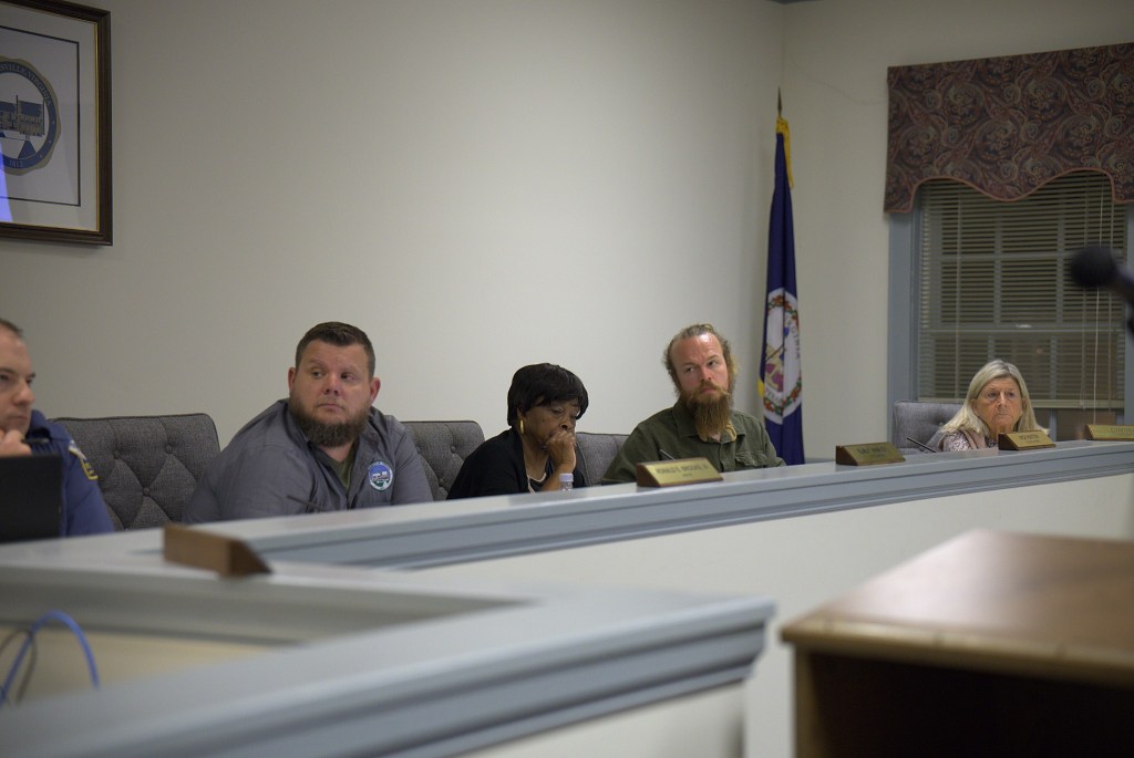 Two men and two women sit behind a white ledge with gray trim in a plain white council meeting room. A window with a paisley valance and a Virginia flag is visible behind them.