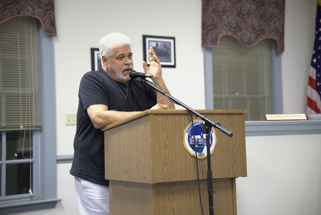 A man in a black tee shirt and white pants leans on a wooden podium with the seal of the Town of Gordonsville on the front and speaks into a microphone. Two windows with purple paisley valances and black and white framed photos are visible on the wall behind him.
