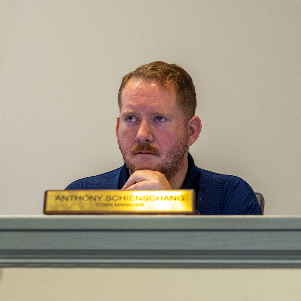 A man in a dark blue polo shirt looks ahead from behind a white ledge with gray trim in a council meeting room. A gold nameplate that reads "Anthony Schienschang Town Manager" rests on the ledge in front of him.