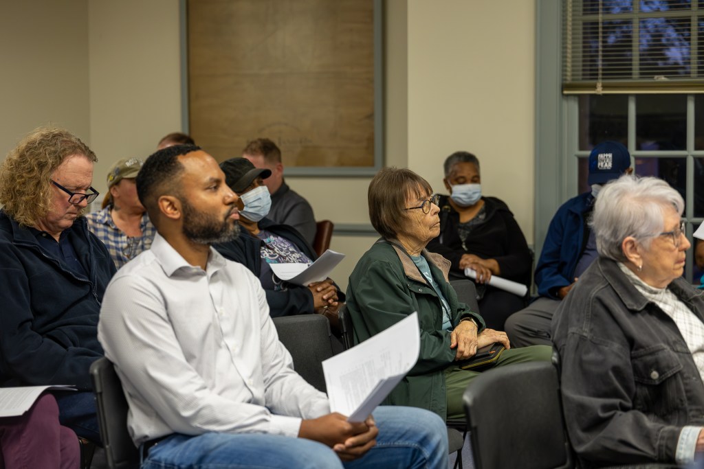A group of people sit in black plastic chairs in a small town meeting room with white walls and gray trim. Some of them are holding papers in their hands, and they are looking forward at someone off-camera.