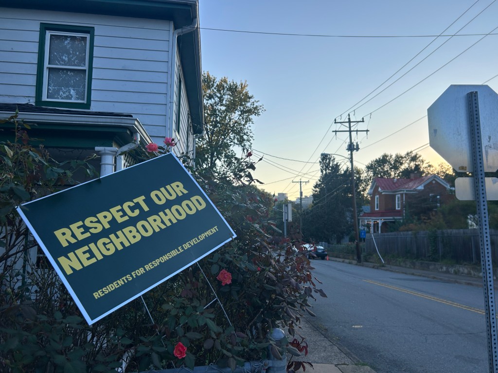 A residential street at dusk. A plastic yard sign sticks out of a rose bush in a front yard in the foreground. The sign reads, in all capital letters, "respect our neighborhood: residents for responsible development." There is a stop sign to the right of the photo, utility lines across the sky, and an older brick home in the background.