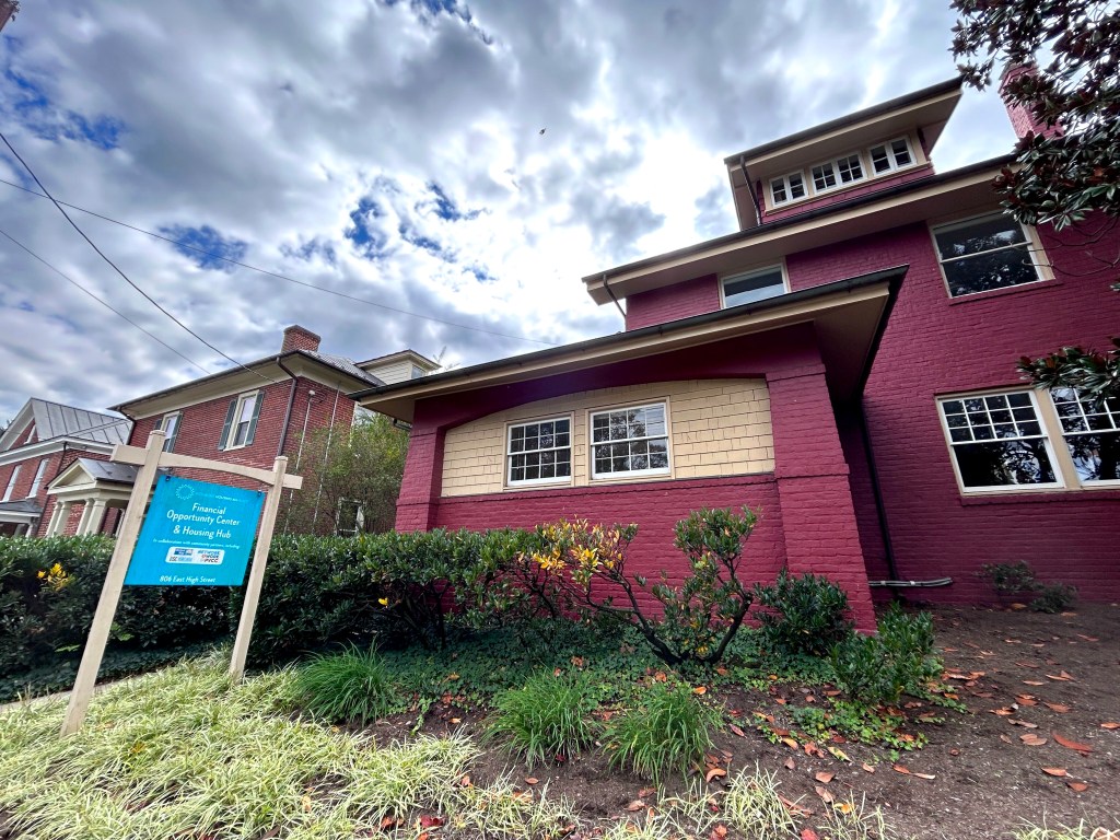A photograph taken of the exterior of a three-story brick and shingle house that now serves as an office. The sky is cloudy, and there are brown and orange leaves among the green plants on the front lawn. There is a sign on the lawn that says "Financial Opportunity Center & Housing Hub." In the background are two similarly-sized, but slightly different style, brick buildings.
