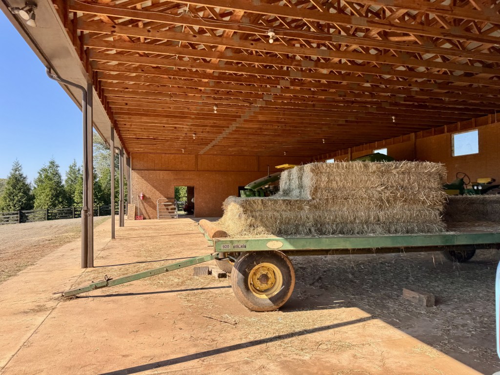 A few square bales of hay are stacked on a green trailer in a long covered barn.