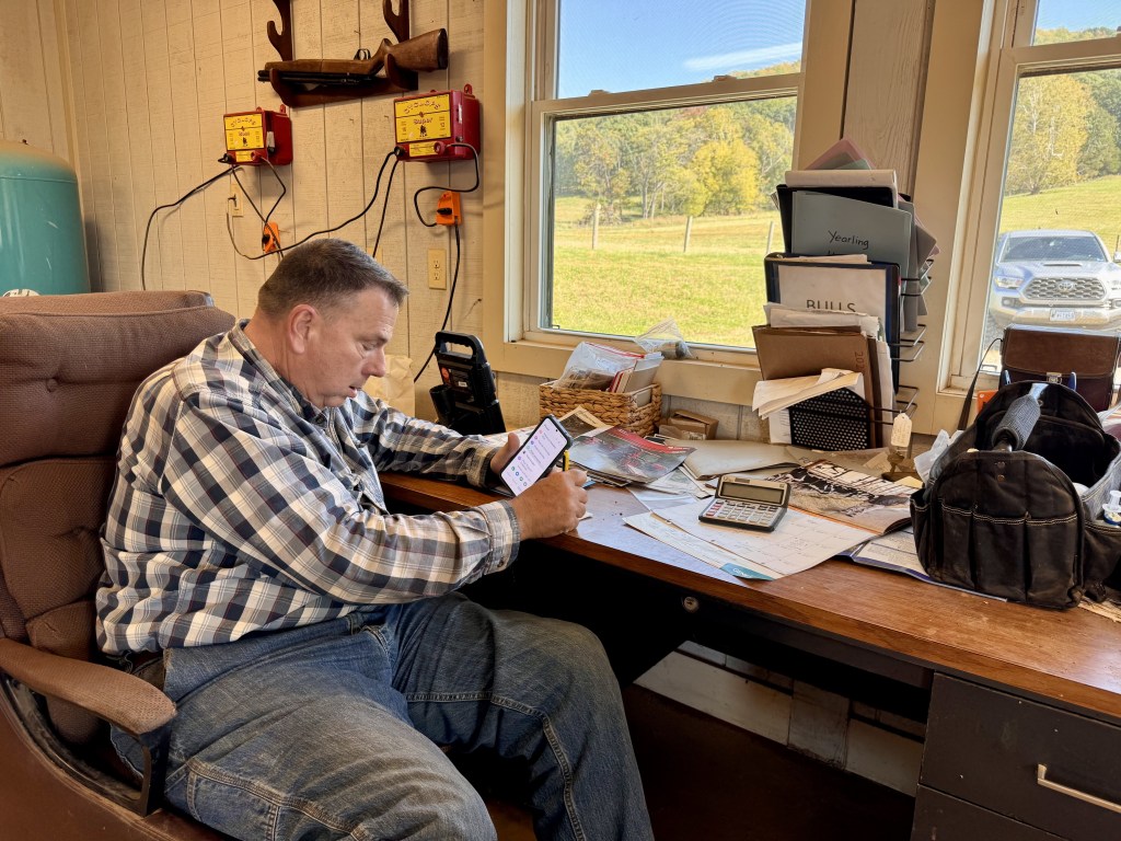 A man in a plaid shirt and jeans sits at a desk with his phone open writing something on a small sheet of paper. The desk is in a older building with wooden walls and sit beneath a window overlooking a pasture and trees.