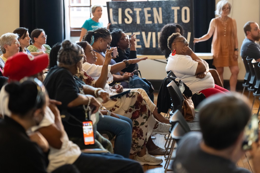 People sit in folding chairs in an auditorium. Two women are animatedly talking and pointing their fingers at someone who is outside of the frame. In the background, two women hold up a banner that says in all capital letters, "listen to Fifeville."