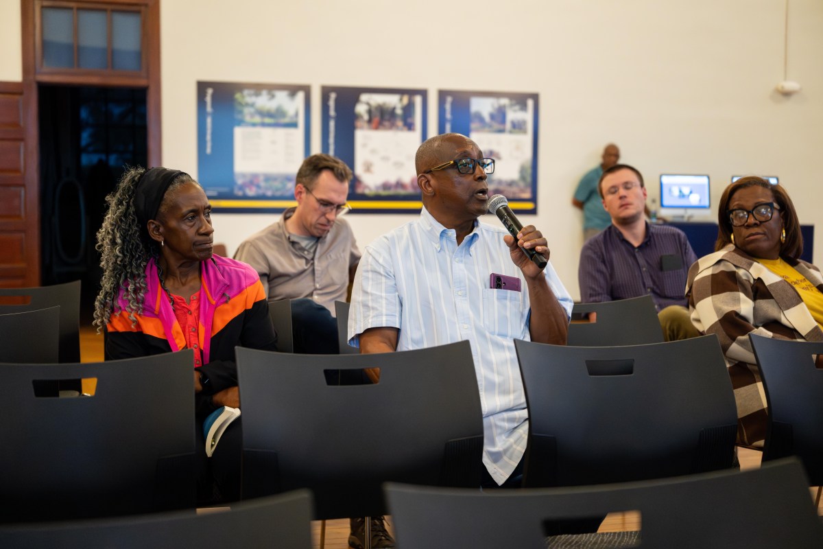 A man speaks into a microphone while seated among other audience members at a community meeting, with informational posters on the wall behind them.