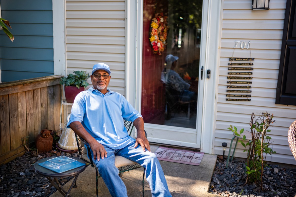 A photograph taken outside, of a Black man sitting in a chair in front of a duplex. He has a gray goatee and wears wire-frame glasses, a blue baseball cap, matching blue polo shirt and light blue jeans. His hands are resting on his thighs, and he looks at the camera, smiling. There is a wreath on the front door of the house behind him, and his reflection is visible in the glass storm door behind him.